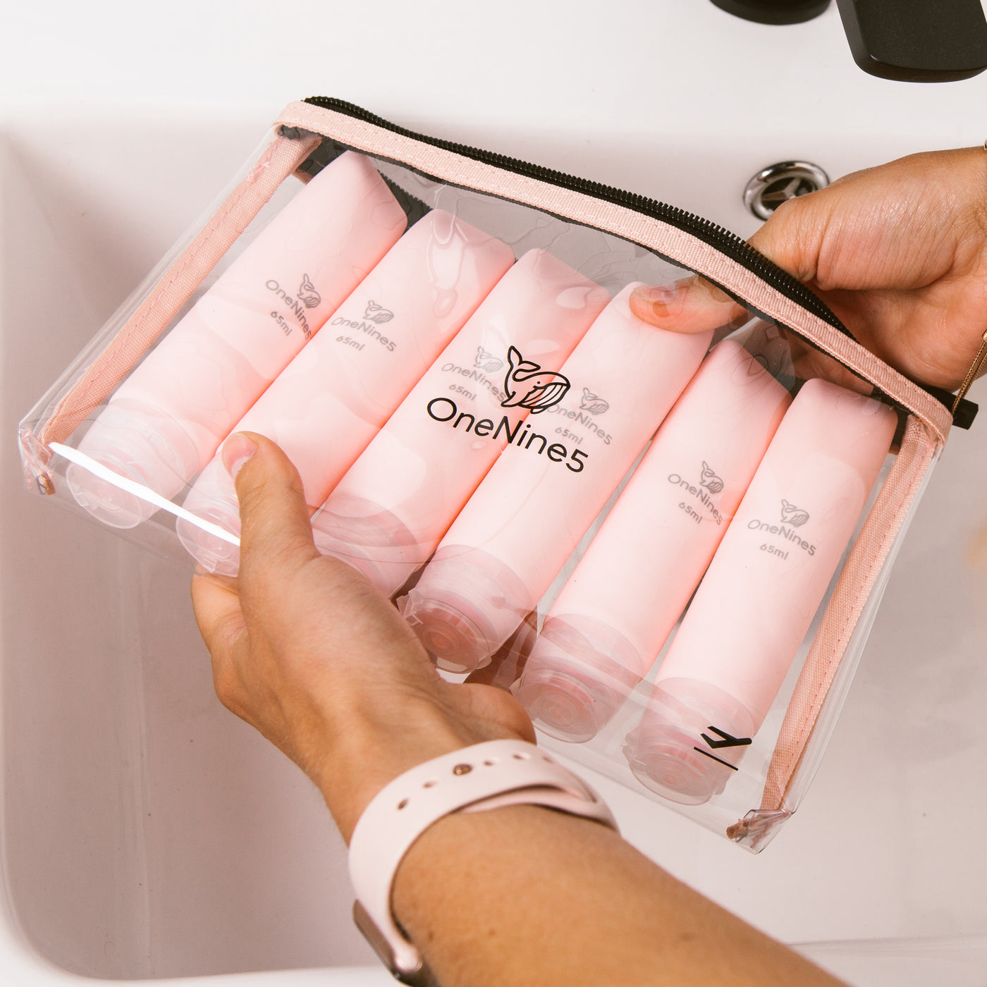 Female hands holding our removable OneNine5 toiletry/ cosmetic clear pouch, with six pink silicone travel bottles packed inside
