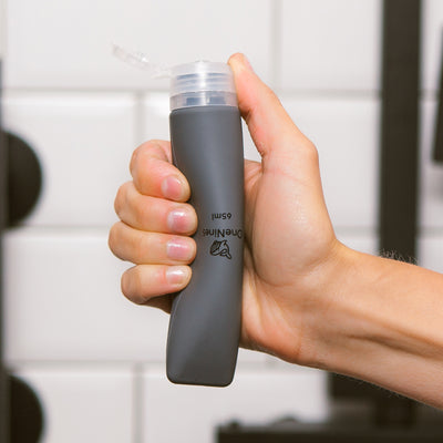 Women's hand squeezing the soft silicone OneNine5 grey travel bottle, with the leakproof cap (lid) flipped open