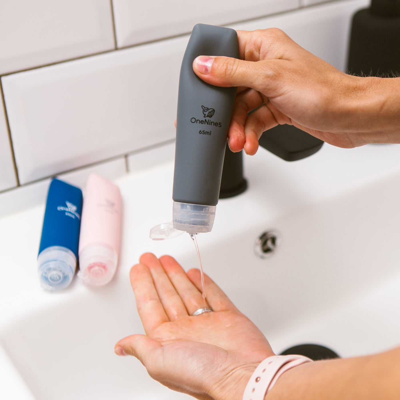 Female squeezing soap from the soft silicone OneNine5 grey travel bottle. Pink and blue reusable OneNine5 bottles are in the background resting on the bathroom sink