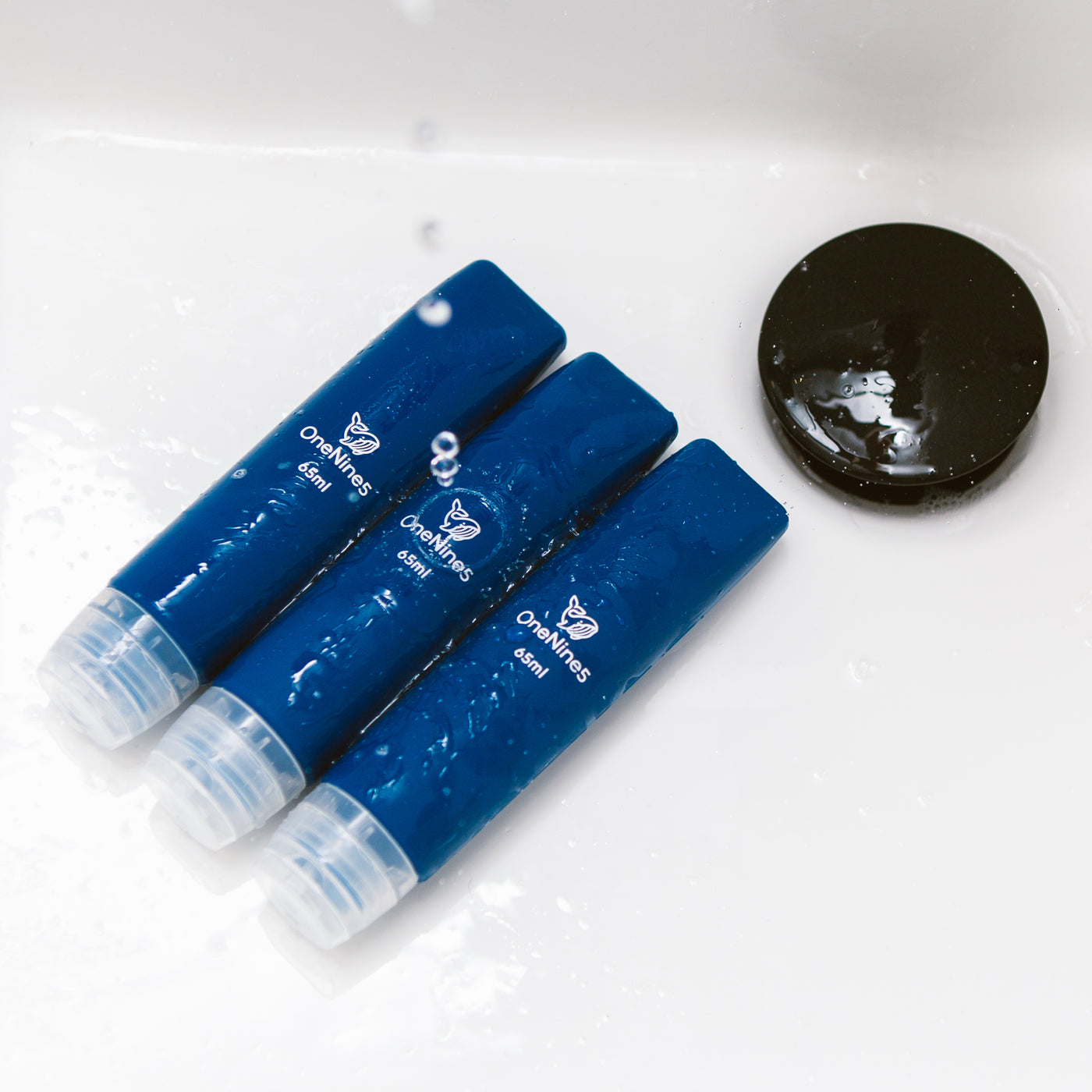 three blue silicone travel bottles in the bathroom sink. A black OneNine5 logo is visible on the reusable bottles and they are being splashed with water