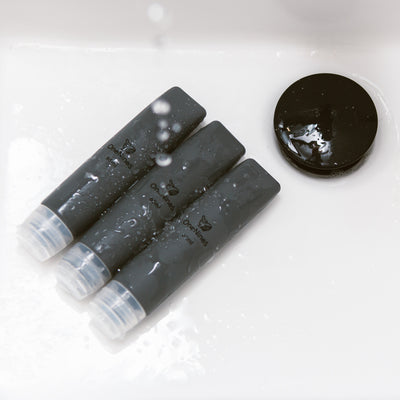 three grey silicone travel bottles in the bathroom sink. A black OneNine5 logo is visible on the reusable bottles and they are being splashed with water