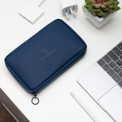 A birdseye view of the OneNine5 Havelock Blue Eco Essentials Pouch, laid flat n a white desk. The debossed OneNine5 logo is visible on the front of the pouch. From the bottom right corner an Apple Macbook and Apple Pencil are visible. At the top of the shot is an Apple USB-C Digital AV Multiport Adapter and a small green plant in a white plant pot.