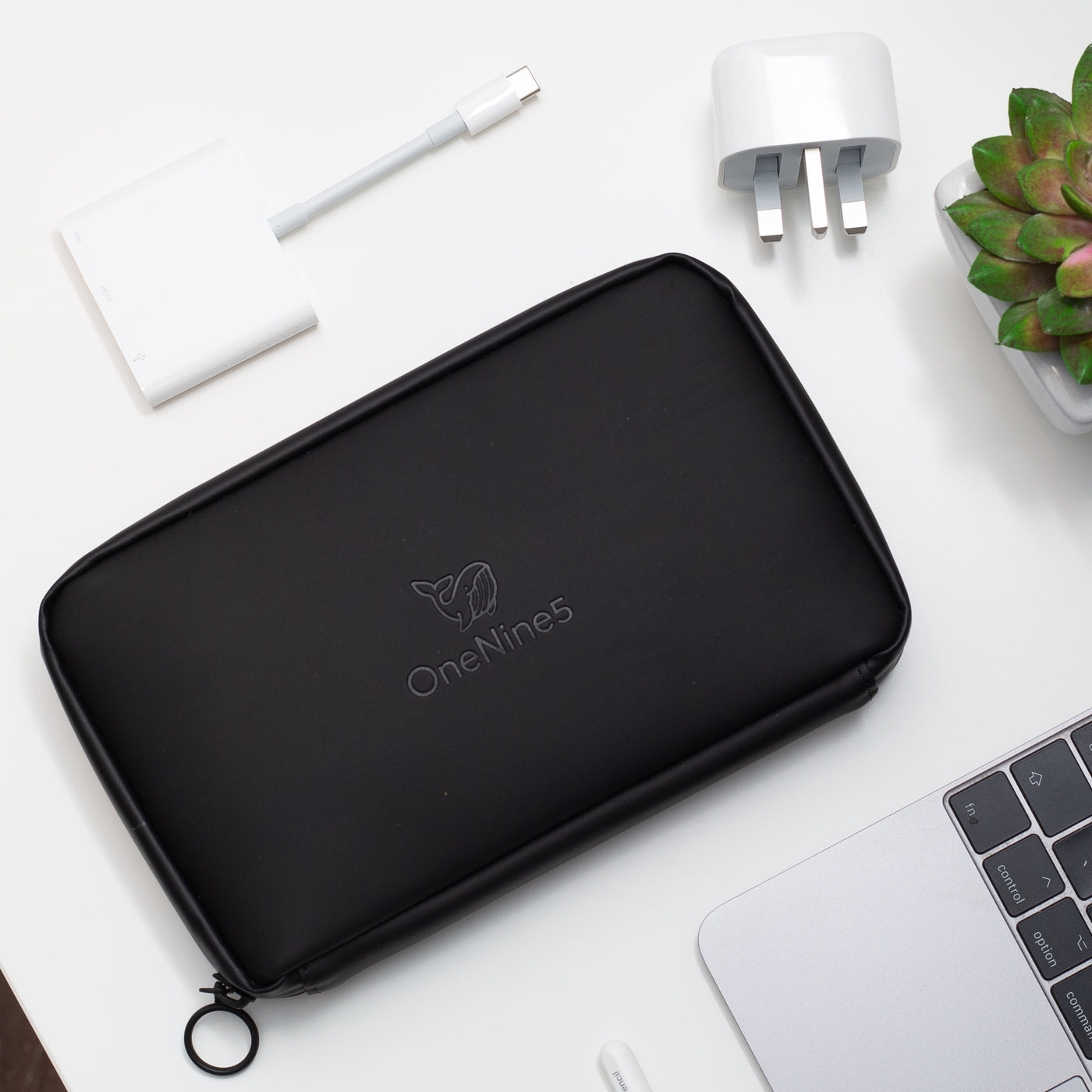A birdseye oview of the OneNine5 Miho Black Eco Essentials Pouch, laid flat n a white desk. The debossed OneNine5 logo is visible on the front of the pouch. From the bottom right corner an Apple Macbook and Apple Pencil are visible. At the top of the shot is an Apple USB-C Digital AV Multiport Adapter and a small green plant in a white plantpot.