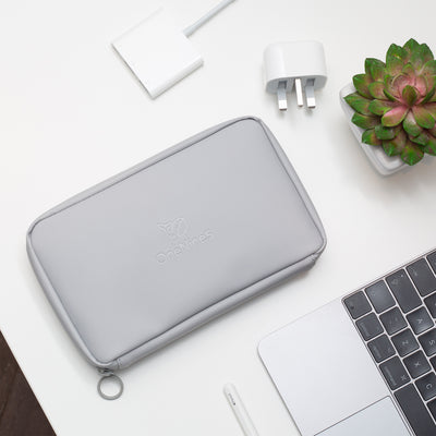 A birdseye oview of the OneNine5 Moeraki Grey Eco Essentials Pouch, laid flat n a white desk. The debossed OneNine5 logo is visible on the front of the pouch. From the bottom right corner an Apple Macbook and Apple Pencil are visible. At the top of the shot is an Apple USB-C Digital AV Multiport Adapter and a small green plant in a white plantpot.