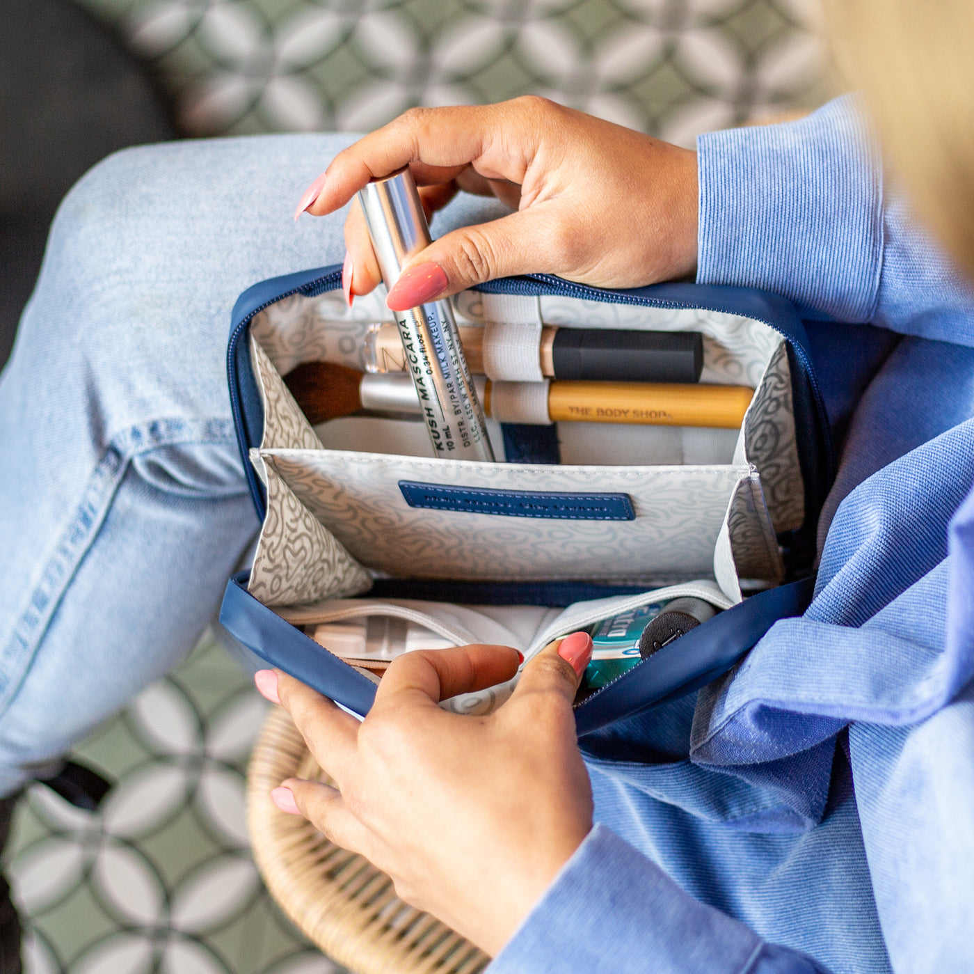 A birdseye view of a women in a blue shirt and blue denim jeans. She's sat with her legs crossed whilst holding open a Havelock Blue Eco Essentials Pouch. Inside the pouch is a range of daily makeup and cosmetics and she's packing Milk Mascara into the bag.