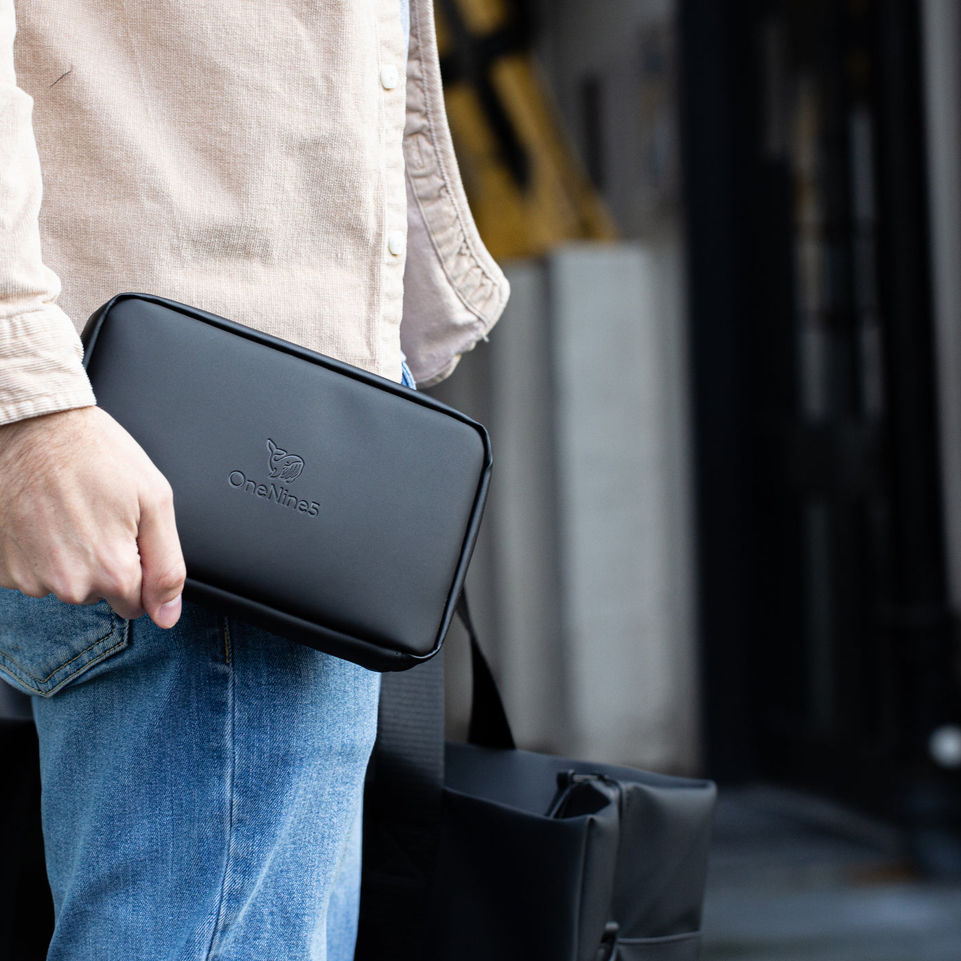 A male on a street wearing blue denim jeans and a beige shirt. Holding a Miho Black Eco Essentials Pouch in his round hand, down by his waist and a weekend holdall bag in his right hand.