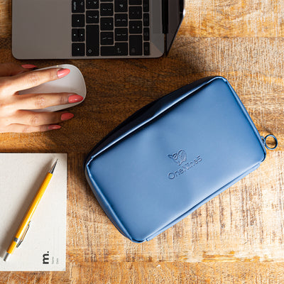A birds eye view of the Havelock Blue Eco Essentials Pouch zipped up and laid flat on a brown wooden desk. A female is working on an Apple Macbook and using an Apple Mouse, with a small grey notebook and and yellow pencil to the left of her