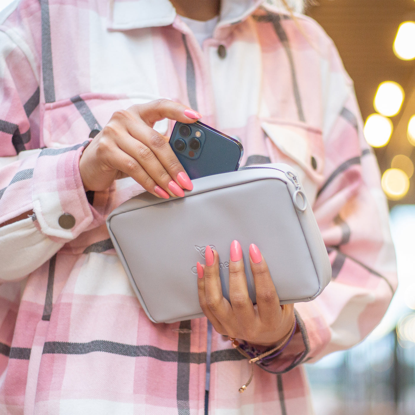 A female wearing a pink cheque shirt. Is holding the Moeraki Grey Eco Essentials Pouch in her left hand. The pouch is partially zipped open and she is placing a blue Apple iPhone 12 pro inside. Her left hand is partially covering the debossed OneNine5 whale logo