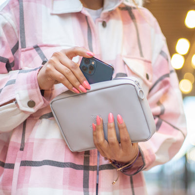 A female wearing a pink cheque shirt. Is holding the Moeraki Grey Eco Essentials Pouch in her left hand. The pouch is partially zipped open and she is placing a blue Apple iPhone 12 pro inside. Her left hand is partially covering the debossed OneNine5 whale logo