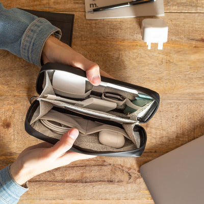 A birdeye view of a male wearing a denim jacket, sat at a brown wooden desk and holding open a Miho Black Eco Essentials Pouch. Inside the pouch is an Apple Watch, Apple Mouse, Apple Airpods, Apple Charging Cable and Apple USB-C Digital AV Multiport Adapter. On the desk is an Apple charging plug, an Apple Macbook, a black pen and a small grey notebook.