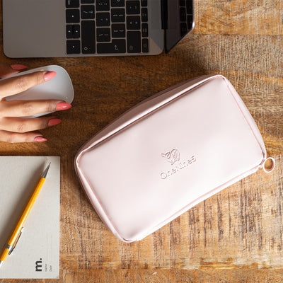 A birdseye view of the Komodo Pink Eco Essentials Pouch zipped up and laid flat on a brown wooden desk. A female is working on an Apple Macbook and using an Apple Mouse, with a small grey notebook and and yellow pencil to the left of her