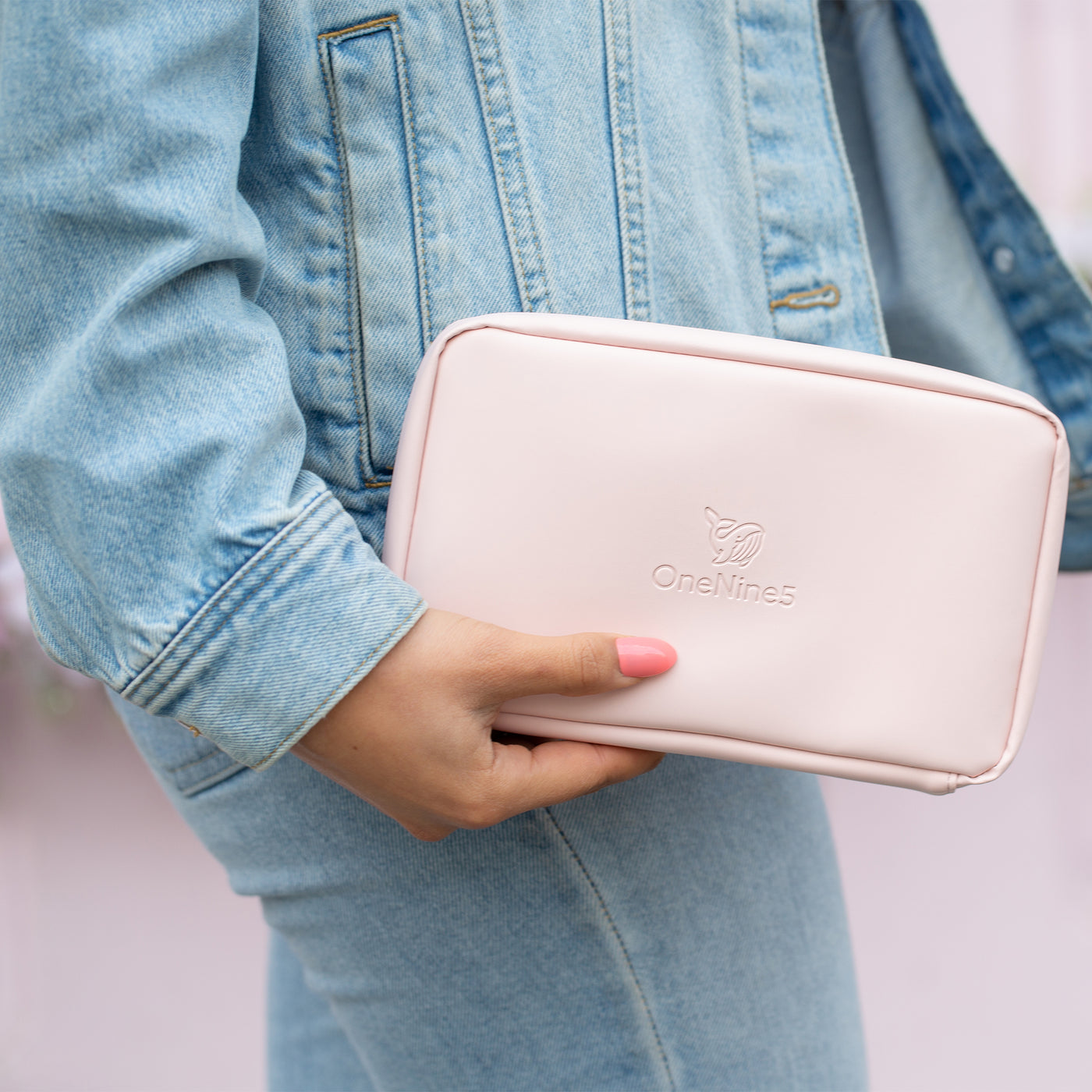 A female wearing a blue denim jacket and blue denim jeans is holding the Komodo Pink Eco Essentials Pouch in her right hand, down by her waist. She is stood in front of a pink shop.