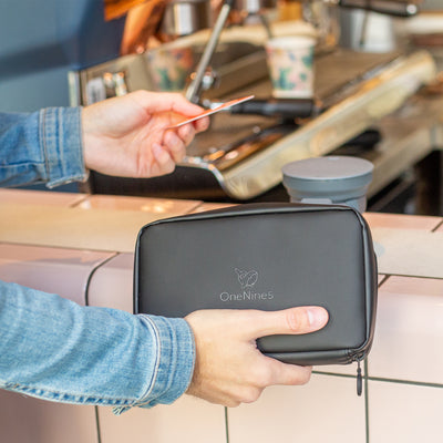 A male in a blue denim jacket is in a coffee shop. He's holding the Miho Black Eco Essentials Pouch and using a Monzo Bank Card to pay for a coffee in his right hand. On the coffee bar in the background is a HUNU coffee cup.