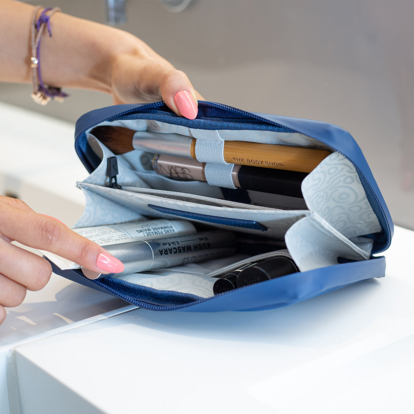 A female in a bathroom packing a range of makeup, cosmetics and toiletries inside a Havelock Blue Eco Essentials Pouch. Inside the range of elastic loops and storage options are visible.