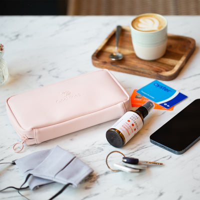 On top of a marble table in a coffee shop, a Komodo Pink Eco Essentials Pouch is zipped up. Alongside the pouch on the table is a face mask, anti-bac spray, a set of keys with a Bluetooth Tile Tracker, an Apple iPhone 12 Pro, an Oyster card and a Monzo Bank Card. In the background is a Flat White on a small wooden tray.