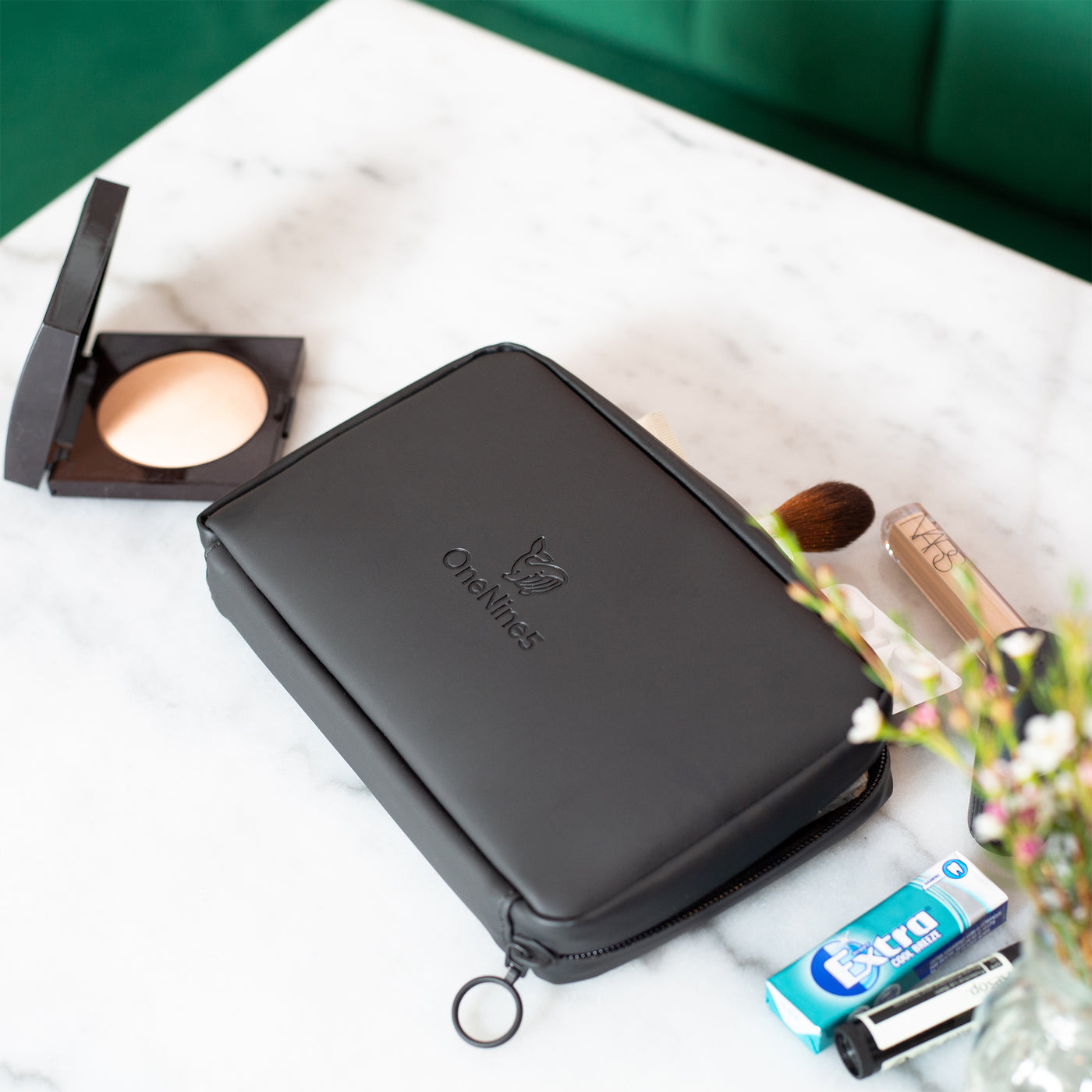 A birdseye shot of the Miho Black Eco Essentials Pouch, laid flat on a marble table with a green velvet bench in the background. On the table alongside the pouch is makeup face powder, NARS Concealer, Wrigleys chewing gum and Aesop lip balm. In the foreground is a slightly blurred out plan in a glass vase.