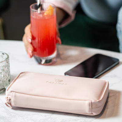 A female in a bar is drinking a brightly coloured red cocktail. In the foreground, she has placed a Komodo Pink Eco Essentials Pouch on the table, next to her Apple iPhone 12 Pro.