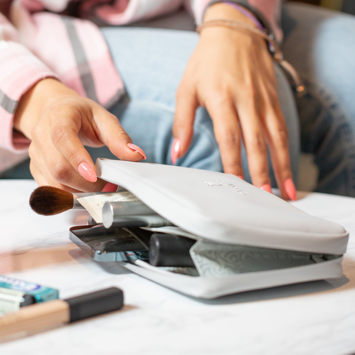 A closeup shot of female hands in the background. The female is wearing blue denim jeans and a pink cheque shirt. In the foreground, a OneNine5 Moeraki Grey Eco Essentials Pouch is zipped open on a white table. A range of makeup and cosmetics are sticking out of the pouch, with NARS Concealer, Wrigleys chewing gum and Aesop Lip Balm in front of the pouch, on the table.